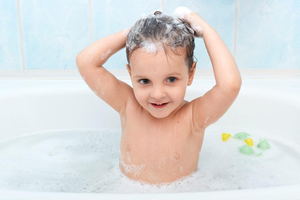 Little girl taking bath, washing her hair with shampoo by herself, looks happy playing with foam bubbles, looking directly at camera, infant enjoys being in warm water, charming baby taking bath.
